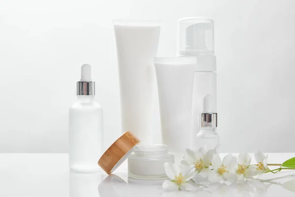 Fleurs de jasmin sur la surface blanche près de bouteilles en verre, crème en tubes, pot et distributeur de cosmétiques — Photo de stock