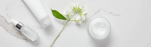 Plano panorámico de jazmín, botella de vidrio cosmético, frasco con crema y tubo hidratante en la superficie blanca - foto de stock