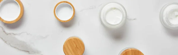 Tiro panorâmico de jarros com nata cosmética e gorros de madeira na superfície branca — Fotografia de Stock