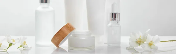 Panoramic shot of cream in tubes, jar and cosmetic glass bottles with jasmine on white surface — Stock Photo