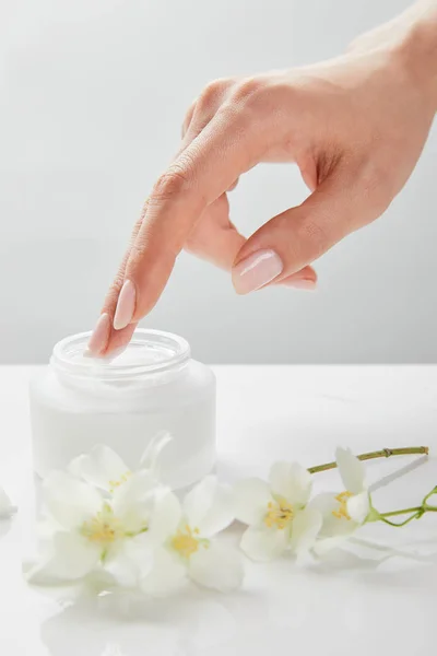 Cropped view of woman hand touching cream in jar near jasmine flowers on white surface — Stock Photo