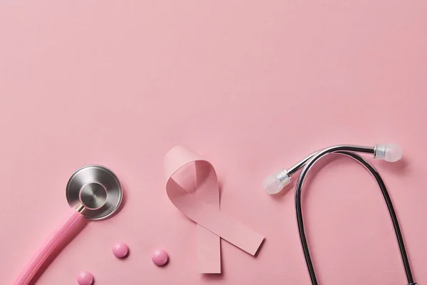 Вид зверху на стетоскоп рожевого металу, стрічка раку молочної залози та три таблетки на світло-рожевому тлі — стокове фото