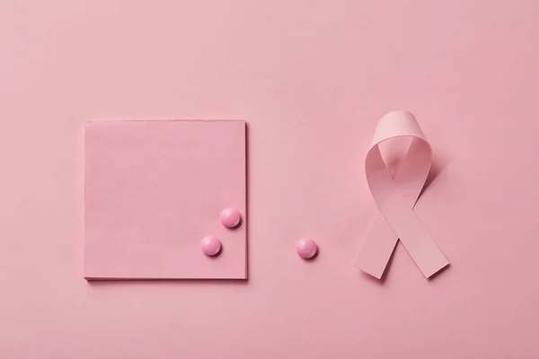 Pilules dispersées sur un morceau de papier près du ruban de cancer du sein sur fond rose clair — Photo de stock