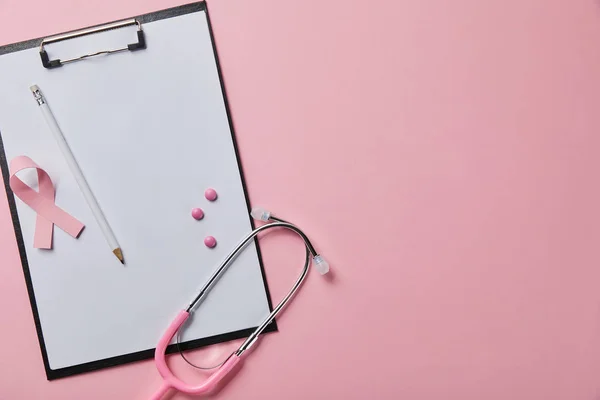 Lápis e fita de câncer de mama na pasta com papel vazio perto do estetoscópio e pílulas espalhadas no fundo rosa — Fotografia de Stock
