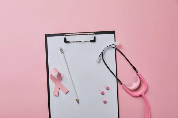 Олівець, рожеві таблетки та стрічки раку молочної залози на папці з порожнім папером біля стетоскопа на рожевому фоні — стокове фото