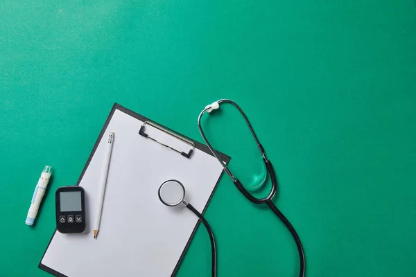 Vista superior de la lanceta de sangre y el estetoscopio cerca del glucosímetro y el lápiz en la carpeta sobre fondo verde - foto de stock