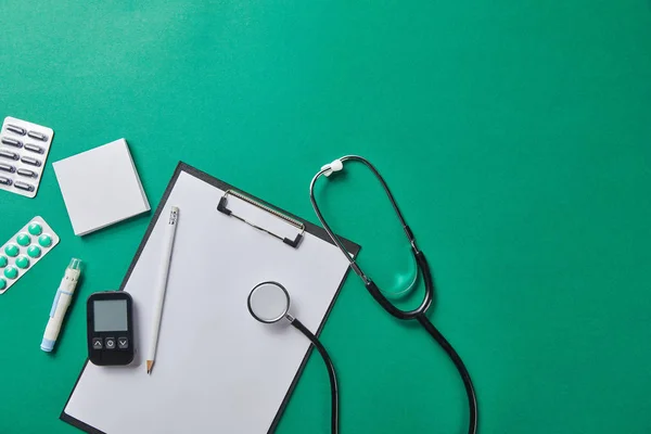 Top view of blood lancet, sticky notes, blister packs and stethoscope near glucometer and pencil on folder on green background — Stock Photo