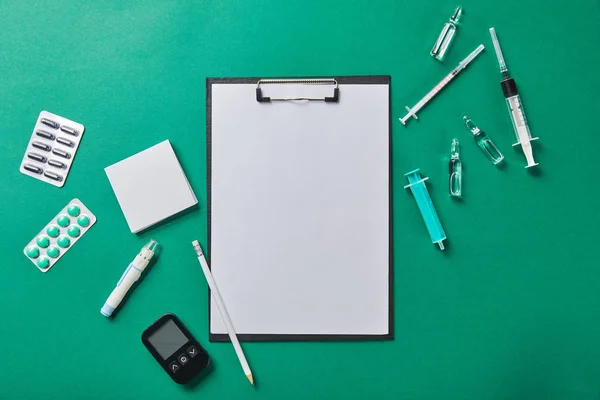 Vista superior del lápiz en la carpeta con papel en blanco redondeado por varios suministros médicos sobre fondo verde - foto de stock