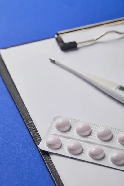 Foyer sélectif de la plaquette thermoformée et du thermomètre sur le dossier avec papier blanc isolé sur bleu — Photo de stock