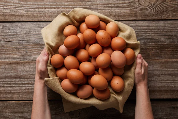 Vista superior del hombre sosteniendo huevos de pollo en tela sobre una mesa de madera - foto de stock