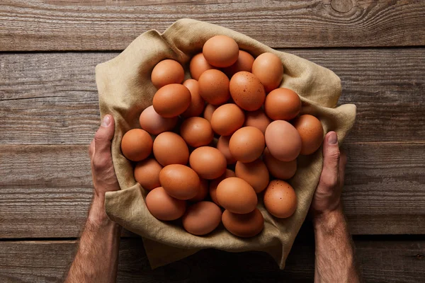 Vista superior del hombre sosteniendo huevos de pollo en tela sobre una mesa de madera - foto de stock