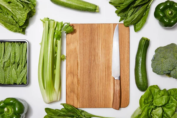 Vista dall'alto di verdure verdi intorno tagliere di legno con coltello su sfondo bianco — Foto stock