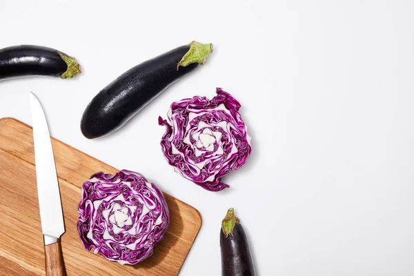 Top view of red cabbage and eggplants on wooden chopping board and knife on white background — Stock Photo