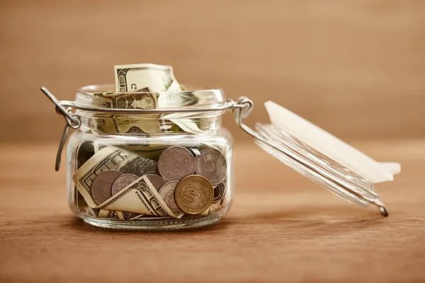 Скляна банка з монетами та доларовими банкнотами на дерев'яному столі — стокове фото