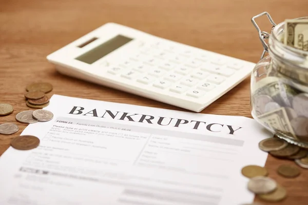 Selective focus of bankruptcy form, coins, calculator and glass jar with dollar banknotes on wooden table — Stock Photo