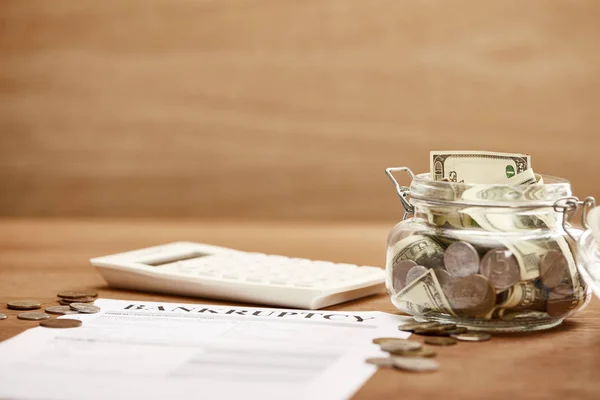 Selective focus of bankruptcy form, coins, calculator and glass jar with dollar banknotes — Stock Photo