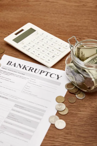 Selective focus of bankruptcy form, coins, calculator and glass jar with dollar banknotes on wooden table — Stock Photo