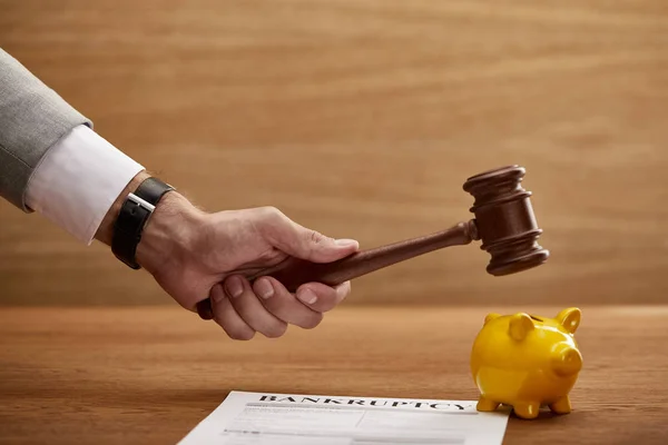 Cropped view of man holding wooden gavel above yellow piggy bank near bankruptcy form — Stock Photo