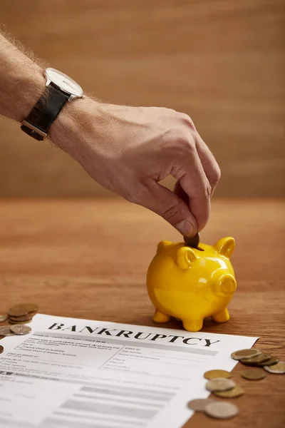 Partial view of man putting coin in yellow piggy bank near bankruptcy form — Stock Photo