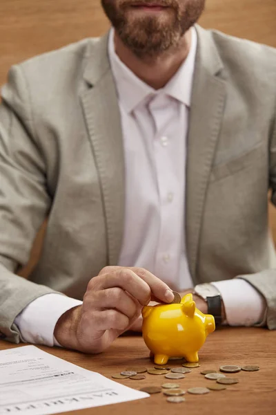 Vista recortada de hombre de negocios en traje poner moneda en alcancía amarilla cerca de la forma de bancarrota - foto de stock