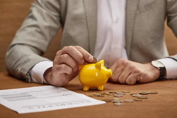 Vista cortada do homem de negócios no terno colocando moeda no banco piggy amarelo na mesa de madeira — Fotografia de Stock