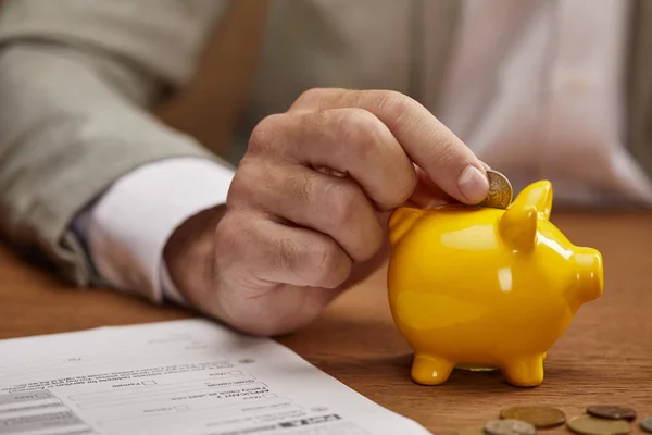 Close up view of businessman putting coin in yellow piggy bank at wooden table — Stock Photo