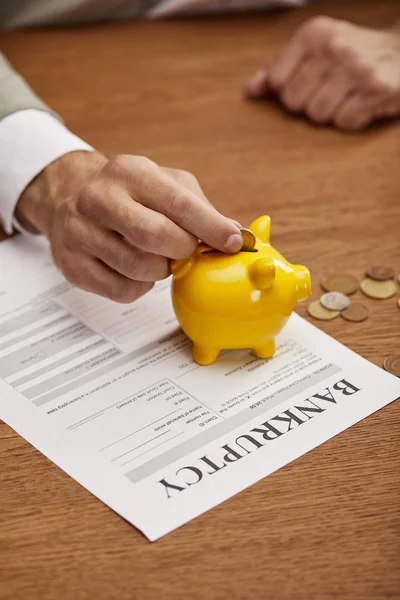 Cropped view of man putting coin in yellow piggy bank on bankruptcy form — Stock Photo
