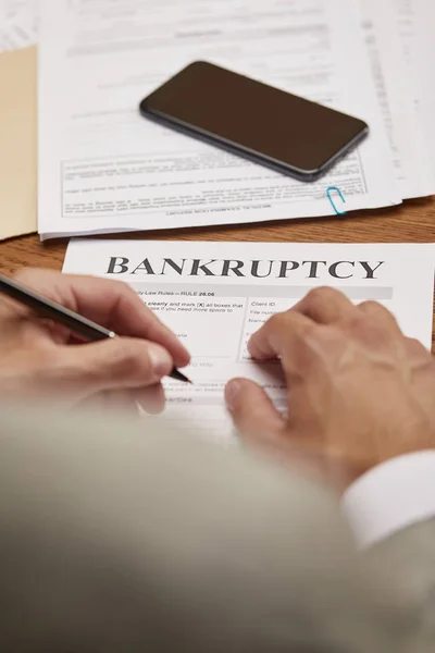 Cropped view of businessman filling in bankruptcy form at wooden table with smartphone — Stock Photo