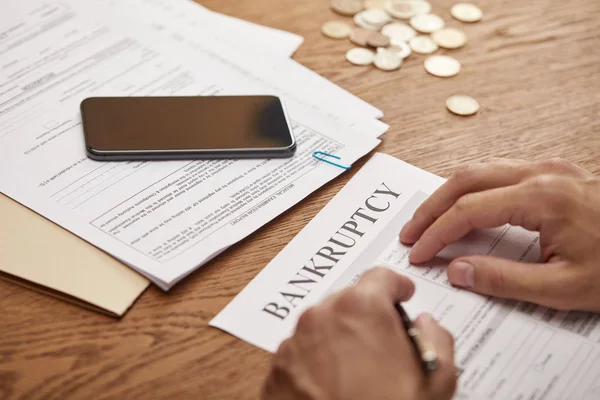 Partial view of businessman filling in bankruptcy form at wooden table with smartphone and coins — Stock Photo