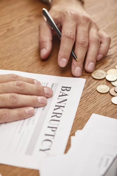 Partial view of businessman filling in bankruptcy form at wooden table with coins — Stock Photo