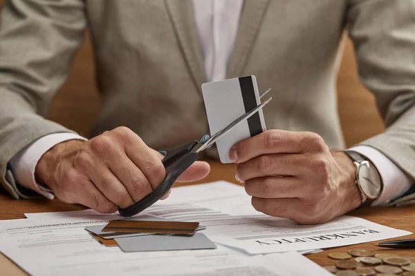 Partial view of businessman in suit cutting credit card with scissors at wooden table — Stock Photo