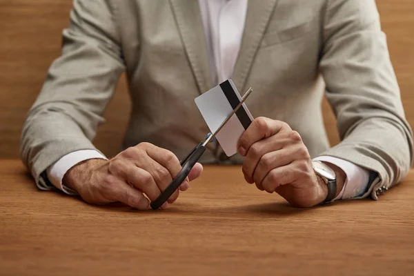 Ausgeschnittene Ansicht eines Geschäftsmannes im Anzug, der Kreditkarte mit der Schere am Holztisch schneidet — Stockfoto