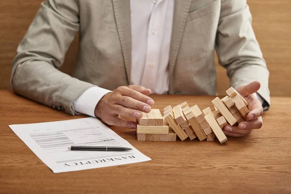 Partial view of businessman in suit holding fallen tower made of wooden blocks near bankruptcy form — Stock Photo