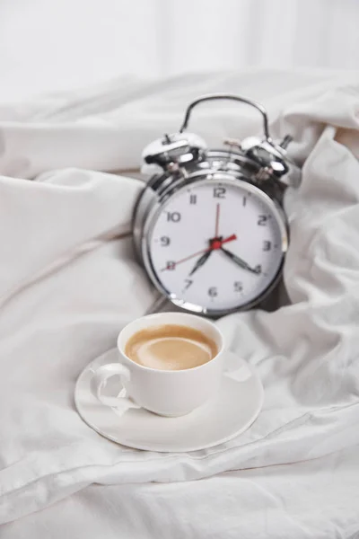 Coffee in white cup on saucer near silver alarm clock in white bedding — Stock Photo