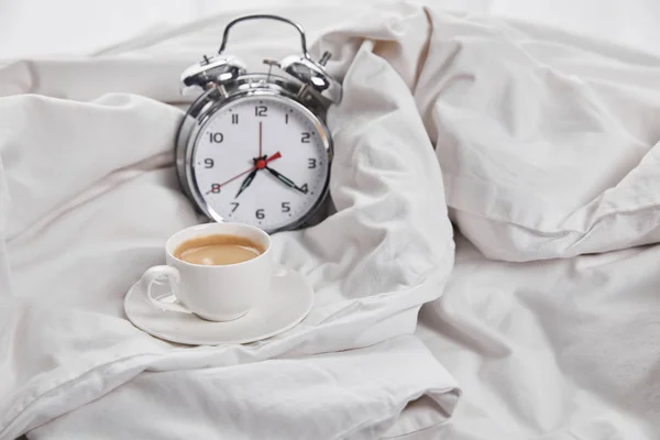 Kaffee in weißer Tasse auf Untertasse in der Nähe von silbernem Wecker im weißen Bett — Stockfoto