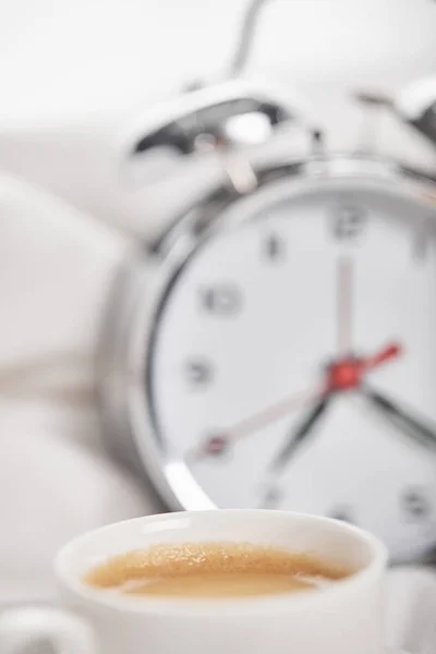 Selective focus of coffee in white cup with silver alarm clock in bed on background — Stock Photo