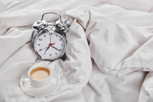 Coffee in white cup on saucer near silver alarm clock in bed — Stock Photo