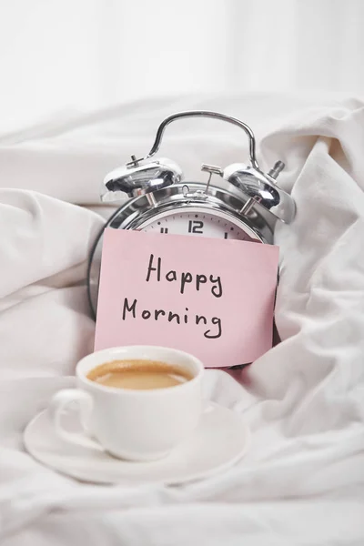 Kaffee in weißer Tasse auf Untertasse in der Nähe von silbernem Wecker mit fröhlichem Schriftzug am Morgen auf klebrigem Zettel im Bett — Stockfoto