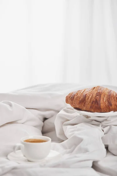 Leckeres Croissant auf Teller in der Nähe von Kaffee in weißer Tasse auf Untertasse im Bett — Stockfoto