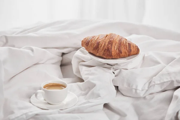 Leckeres Croissant auf Teller neben Kaffee in weißer Tasse auf Untertasse im Bett — Stockfoto