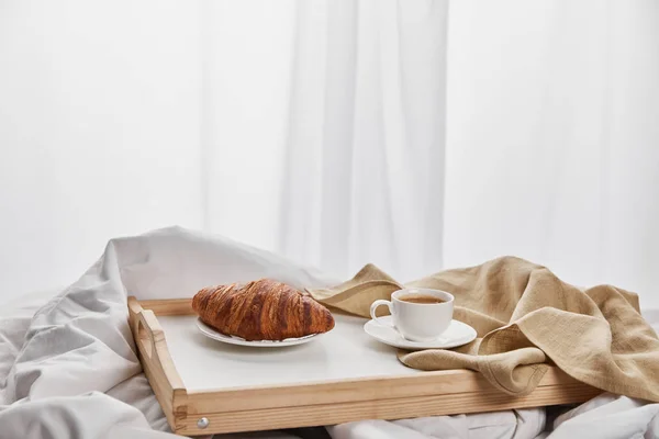 Frisches Croissant mit Kaffee in Tasse auf Holztablett im Bett am Morgen — Stockfoto