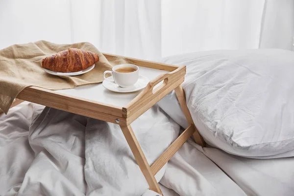 Frisches Croissant mit Kaffee auf Holztablett im Bett mit Kopfkissen am Morgen — Stockfoto
