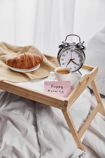 Серебряные будильники, липкая нота со счастливым утренним надписью, кофе и круассан на деревянном подносе на белом постельном белье — стоковое фото