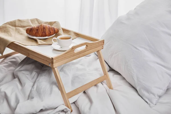 Café y croissant servido en bandeja de madera sobre ropa de cama blanca - foto de stock