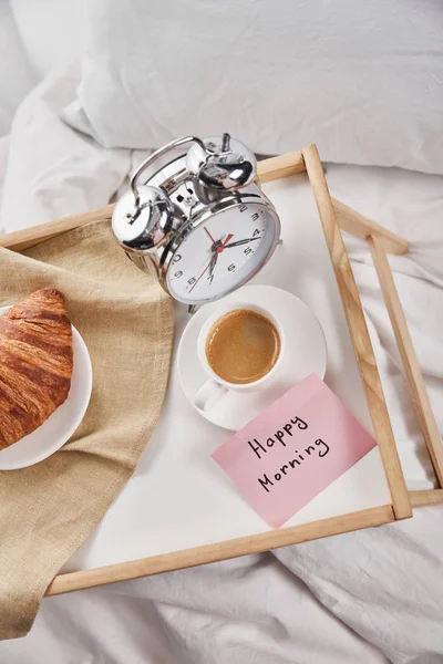 Top view of alarm clock, sticky note with happy morning lettering, coffee and croissant on wooden tray on white bedding — Stock Photo