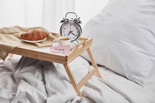 Despertador, nota adhesiva con letras de la mañana feliz, café y croissant en bandeja de madera sobre ropa de cama blanca - foto de stock