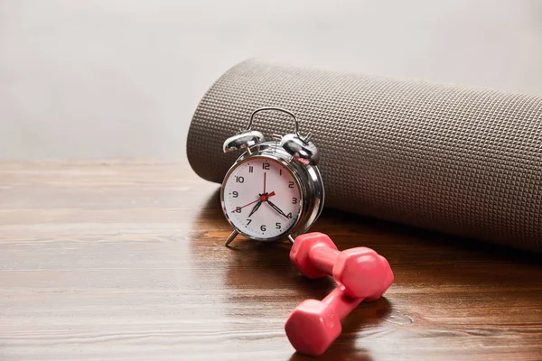 Silver alarm clock near dumbbells and fitness mat on wooden table isolated on grey — Stock Photo