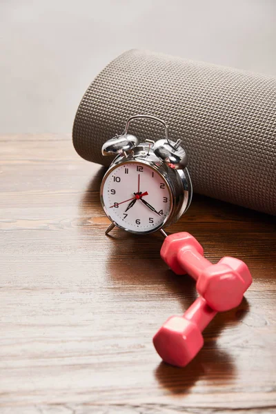 Despertador plateado cerca de pesas rosadas y colchoneta de fitness en mesa de madera aislada en gris - foto de stock