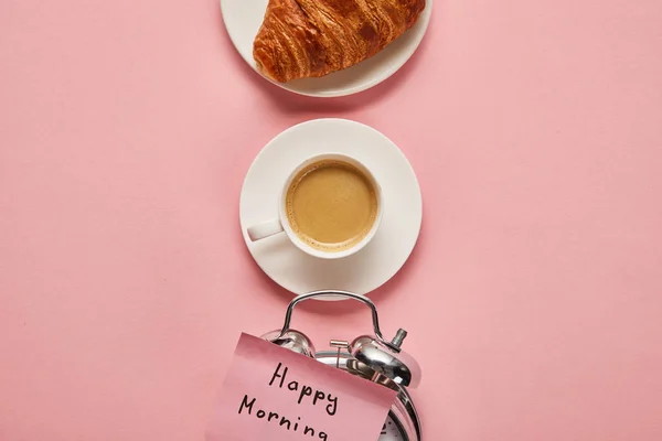 Flache Liege mit Wecker mit Happy Morning Schriftzug auf klebrigem Zettel in der Nähe von Kaffee und Croissant auf rosa Hintergrund — Stockfoto