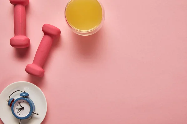 Вид сверху на апельсиновый сок, розовые гантели и игрушечные будильники на тарелке на розовом фоне — стоковое фото